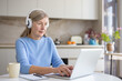 © Liubomir - Senior woman concentrating on her laptop screen, wearing white headphones while typing on the keyboard and sitting at a round white table in a bright modern kitchen
