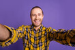 © deagreez - Smiling young handsome guy in casual checkered plaid shirt takes a selfie against a purple background showcasing fashion model