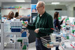 © DC Studio - Retired male person browses vitamins and wellness products in drugstore, reading medicine packages labels to highlight professional pharmaceutical care. Modern dispensary setting.