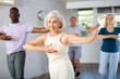 © JackF - Elderly woman is engaged in dance lesson and repeats basic movements for pair ballroom dancing. Amateurs beginners of different ages clients of fitness studio dance in background