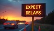 © Maryna - Electronic traffic warning sign shows EXPECT DELAYS message on highway at sunset. Blurred cars pass by road cones on road side. Road work congestion causes traffic hold up for drivers. People face