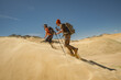 © ANDREY - A young couple with backpacks ascends a sand dune, kicking up a small sandstorm with their feet. Side view shows them moving right to left during their desert trek.