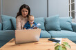 © Srdjan - Working mom with baby on lap reviews household bills on a laptop at home balancing childcare and finances; determined to solve budgeting issues while managing payments subscriptions and savings goals