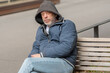 © Anelo - An elderly man in a hoodie is sitting on a city bench, looking tired and thoughtful.