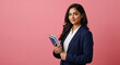 © PRASANNAPIX - A confident young Indian woman in a navy blazer holds a notebook and pencil, standing poised against a pink background,