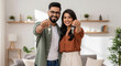 © PRASANNAPIX - A young Indian couple stands together in a modern living room, smiling and proudly holding keys to their new home.