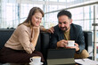© pressmaster - Caucasian young adult woman and Middle Eastern young adult man sitting together at table in car dealership discussing information on digital tablet while drinking coffee