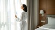 © PurArts Studio - hotel concept Woman in white bathrobe opening curtains in a hotel room to greet the morning sun