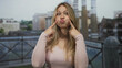 © Krakenimages.com - Blonde woman wearing a pink top points finger to puffed cheeks against rome street railing with blurred ruins; playfulness humor silliness.