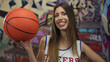 © Krakenimages.com - Cheerleader smiling and holding basketball on urban street with graffiti wall; team spirit confidence.