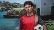 © Krakenimages.com - Man holding soccer ball at a seaside building wearing a red team jersey and smiling by the coast; pride.