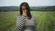 © Krakenimages.com - Woman in striped shirt touching eyeglasses with two fingers while standing among vineyard rows in a forest; quiet thoughtfulness.