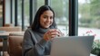 © mahamudul - Young woman smiling and holding a coffee cup while working on a laptop in a cozy cafe
