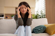 © Dexon Dee - A woman sitting on a sofa with her hands on her temples, looking sad and stressed as if dealing with heartbreak, job loss, or financial worries at home.