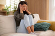 © Dexon Dee - A woman sitting on a sofa with her hands on her head, looking distressed and overwhelmed while experiencing emotional stress at home.
