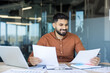 © Liubomir - Young businessman analyzing reports and documents at his office desk, confidently reviewing financial data and smiling, showing success and professional growth in a modern workspace