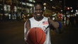 © Krakenimages.com - Man smiling holds basketball and points finger to ball on street at night with city lights and buildings; urban joy.