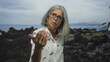© Krakenimages.com - Woman extends open hand toward viewer on rocky seaside by a building, glasses visible and expressive face; pleading.