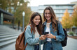 © Vadym - Two girls look at phone screen, laugh in campus. Female students watch funny content. Young friends use modern mobile. Girlfriends hold smartphone outdoors. People smile on university street enjoy