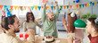 © Studio Romantic - Joyful man raising hands while celebrating birthday with friends. Group of happy diverse people at desk wearing party hats cheering, clapping, enjoying cake and gifts in decorated living room. Banner.