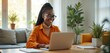 © Viktor - Young Black woman smiles working on laptop at home desk. She uses computer for remote job, online research or social media posts. Modern apartment with plants and window light.