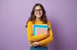 © Viktor - Young female student smiles, holds colorful books. Wears glasses, stands against purple wall. Girl looks happy, ready for learning, back to school season. Future education success, university life,