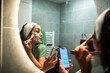 © Marko Geber - Young adult woman applying face mask and texting in home bathroom, playful