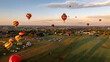 © Greg Kelton - Hot air balloons of various colors soar in the evening sky during a festival in the countryside. The landscape is vibrant with green fields and distant hills under a warm sunset.