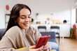 © Xavier Lorenzo - Happy young woman using mobile phone sitting on sofa at home, enjoying cozy moment chatting with friends or watching social media content online. Technology lifestyle concept