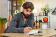 © Andrii Iemelianenko - Indian man at home listens to audiobook with headphones takes notes in notebook stays focused. Arabian guy at desk writes key ideas marks quotes learns skills keeps attention and reviews notes after