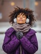© Johannes - afro american woman feels very cold during freezing weather wears purple jacket and warm scarf around neck wals on street during winter listens music via wireless headphones keeps hands together no l