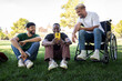 © Daniel - African American man in a wheelchair and friends sitting on the grass looking at mobile phone in a park.