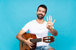 © luismolinero - Young man with guitar over isolated blue background happy and counting four with fingers