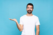 © luismolinero - Young man with beard  over isolated blue background holding copyspace imaginary on the palm to insert an ad