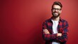 © miss irine - Young man with beard and glasses smiles confidently. He wears a plaid shirt over a white tee, arms crossed against a red backdrop, looking directly ahead.