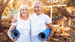 © Prostock-studio - A happy senior couple stands smiling while holding their yoga mats in a bright and sunny park. The trees have autumn leaves, and they look ready for their yoga session together.