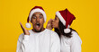 © Prostock-studio - A man reacts with surprise while a woman whispers in his ear, both wearing festive Santa hats. The bright yellow background adds to the joyful holiday atmosphere.