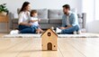 © lamin - Wooden house toy on living room floor while smiling family plays behind, showing home buying process, mortgage planning, property security, rental choice, children and housing stability