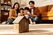 © lamin - Small wooden house model on living room floor with joyful family in background, showing real estate ownership, home loan planning, housing investment, rental options, children and future happiness