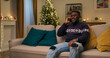 © ABCreative - A young student sits on a sofa, having received a Merry Christmas call from a friend. He is very happy to communicate and smile, wearing a warm sweater and ripped jeans.