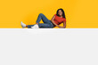© Prostock-studio - A young woman lies comfortably on a white surface wearing a red shirt and blue jeans. She smiles while resting her head on one hand, with a vibrant yellow background enhancing the cheerful mood.