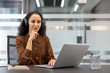 © Liubomir - Young woman with curly hair wearing headphones and an earth tone shirt. Concentrating while using a laptop from a desk in a contemporary co-working office. Focusing on an online webinar or remote work