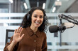 © Liubomir - Young female broadcaster wearing headphones and speaking into a microphone, greeting the audience while recording or streaming content in a modern studio environment