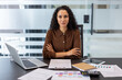 © Liubomir - Confident businesswoman with arms crossed at her desk, serious and focused amid laptop, documents and financial charts, conveying professionalism, leadership and corporate success in office