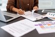 © Liubomir - Person signing a legal business contract or agreement document with a pen, sitting at an office desk with other paperwork, laptop, and charts during work