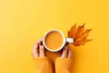Mains féminines tenant une tasse de café sur fond jaune uni avec une feuille d’automne et espace négatif copyspace, visuel chaleureux pour la Journée mondiale du café célébrée le 1er octobre