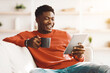 © Prostock-studio - A young man in a red sweater sits comfortably on a couch, smiling as he reads on his tablet and holds a cup of coffee in a bright and inviting living room setting.