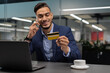 © Prostock-studio - Young businessman is smiling while holding a credit card in one hand and speaking on a mobile phone. He appears satisfied after making an online purchase in a modern office setting.