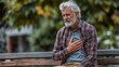 © Bella - Middle-aged man with gray hair, sitting outdoors and clutching his chest, experiencing sharp pain, a potential sign of a heart attack or cardiac distress.