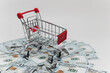 © vladdeep - A miniature shopping cart rests atop a pile of US hundred-dollar bills against white background